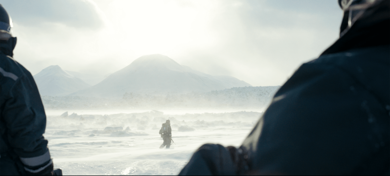 Plan large, un homme marche dans le blizzard face à un glacier, au premier plan deux personnages le regardent