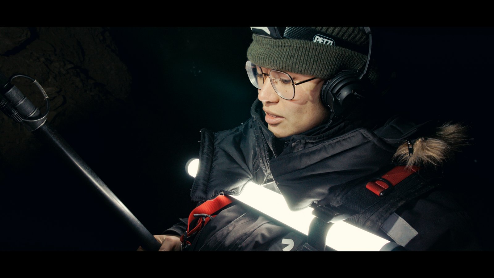 Plan large, une jeune femme avec des lunettes et un casque audio tient une perche et une lumière