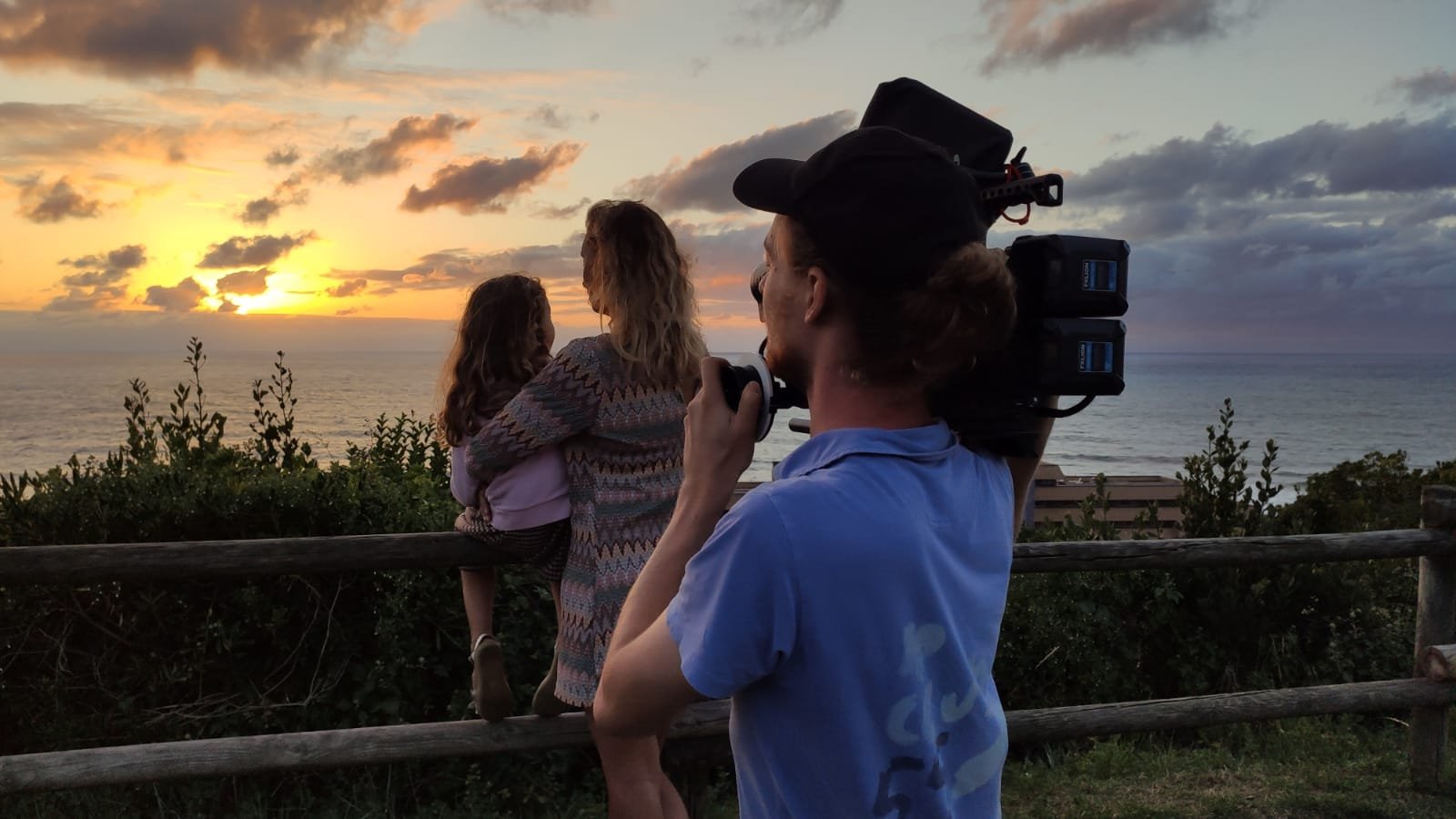 Plan large, un homme filme une femme et sa fille devant un coucher de soleil